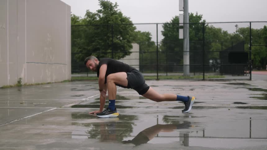 A man performs leg and arm stretches on a wet tennis court