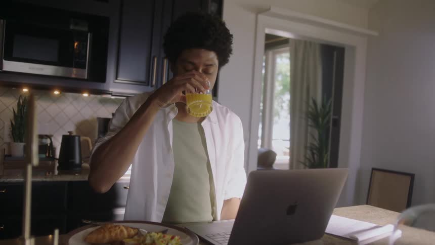 A man is sitting looking at a laptop that is on the table and he is drinking something from a drink glass