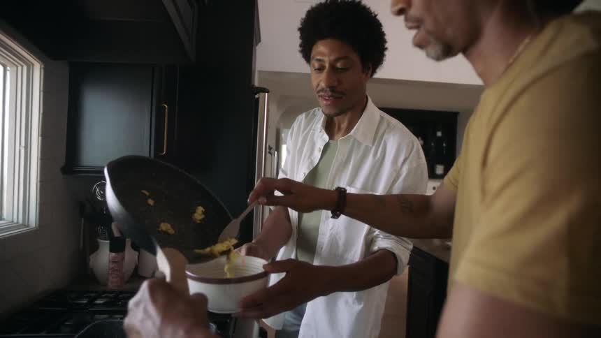 Two men in a kitchen one holding a pan from which food is being served into a bowl