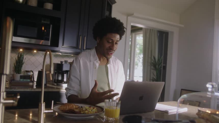 Man sitting in the kitchen talking with a laptop in front of him and a plate of food with a glass of juice next to him