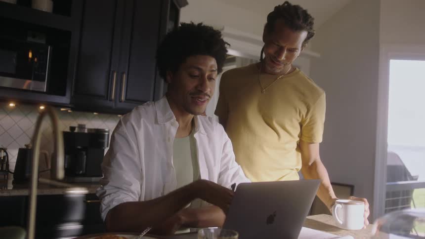 Two men in the kitchen with one of them pouring a cup and the other working on a laptop