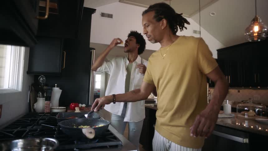 Two men in a kitchen cooking with frying pans and one of them tasting