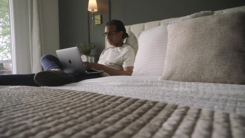 A black man sits on his bed while typing on a laptop