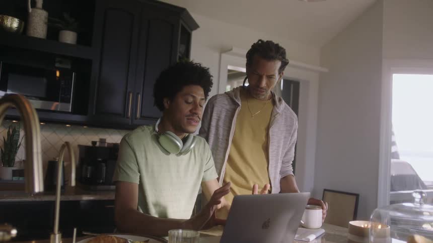 Two men are talking and looking at a laptop that is on a table in a kitchen