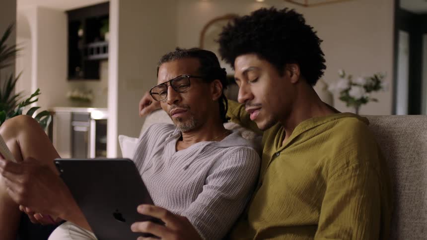 Two men sitting together hugging each other with one of them reading a book and the other using a tablet