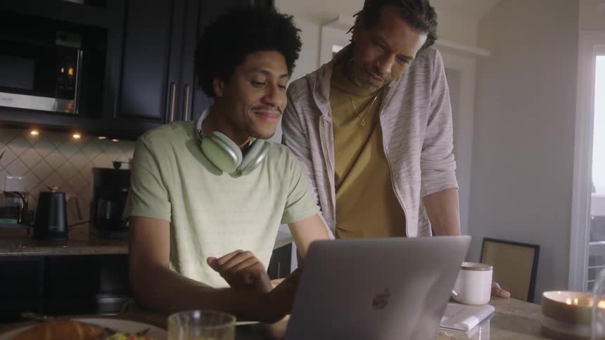 A couple in the kitchen one seated at a counter using a laptop the other standing beside him