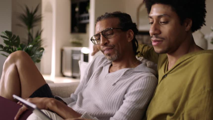 Couple of two men sitting on the sofa with one of them reading a book and the other using a tablet