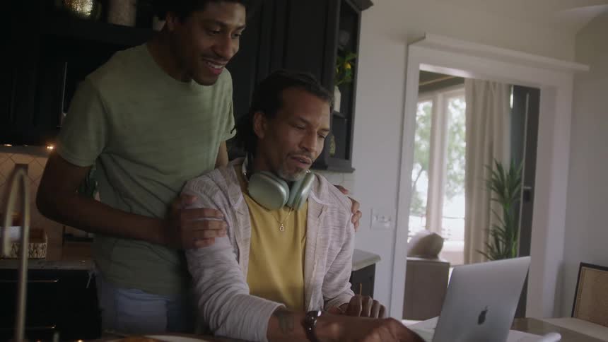 Two men in the kitchen with one of them wearing headphones and sitting on his laptop