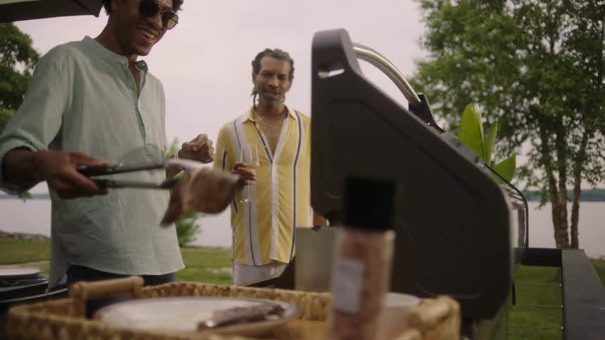 A black man cooks on a grill while his friend drinks wine and watches