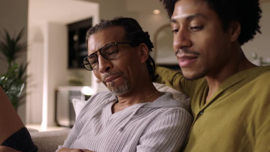 Two men sitting on a sofa reading a book with one of them holding a tablet