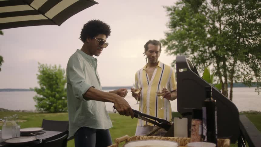 Man preparing barbecue with his colleague next to him holding two glasses of wine
