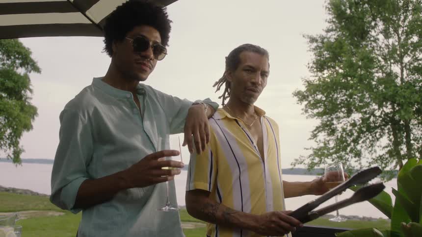 Two men in front of a barbecue and holding glasses of wine