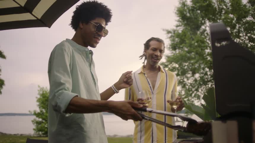 Two men preparing a barbecue with one of them holding glasses of white wine