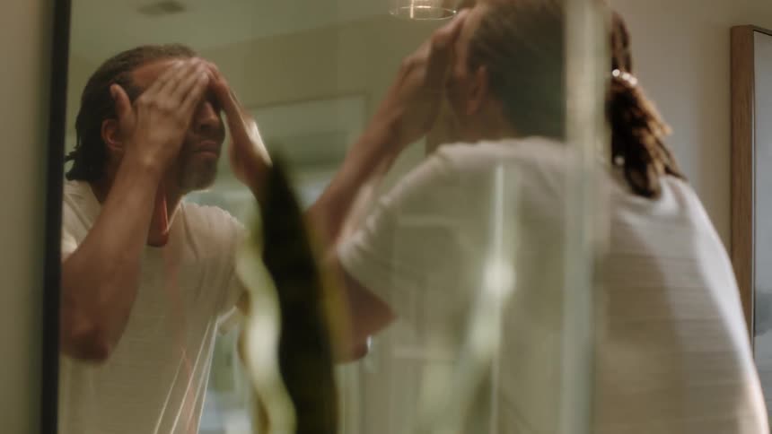 A black man rubs his face while standing in front of a mirror