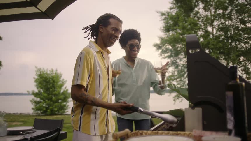 A man flips steaks on a barbecue while his friend holds two glasses of wine