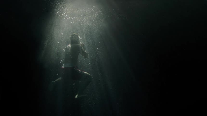 A white males emerges from the depths of a body of water and swims up to the light