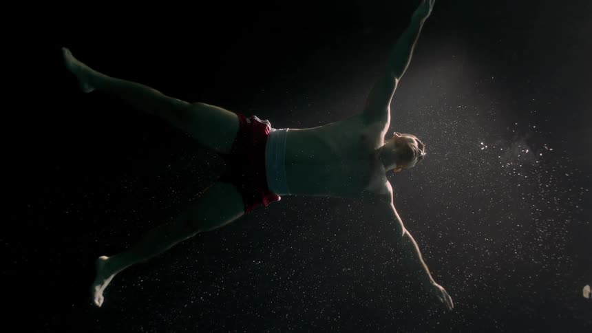 A man is underwater and shirtless and swimming to the surface
