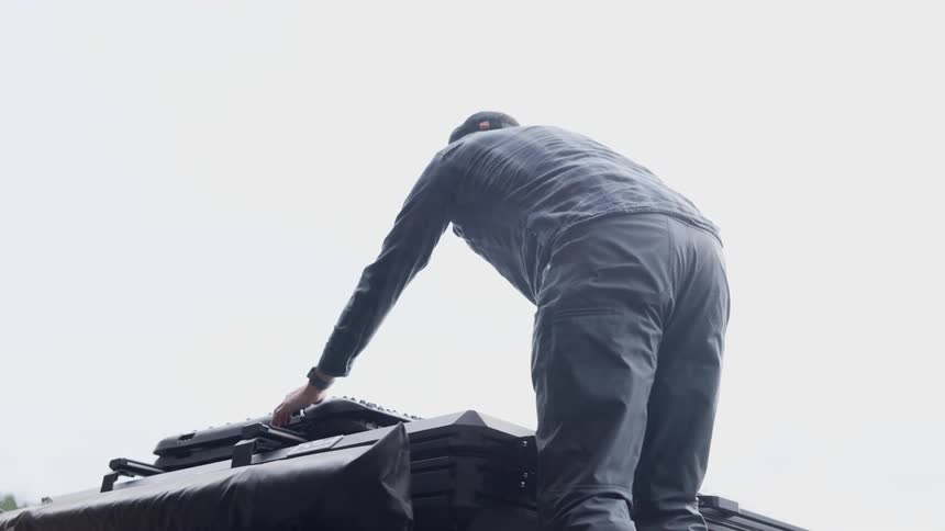 A man climbs on top of a vehicle to make adjustments to an object strapped on the roof