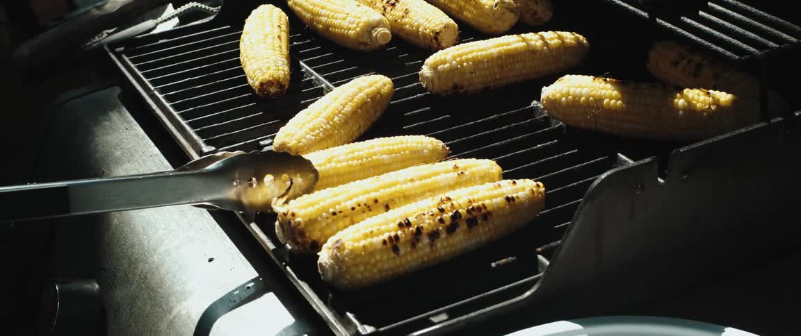 Person with a metal spatula turning corn that is being roasted on a barbecue