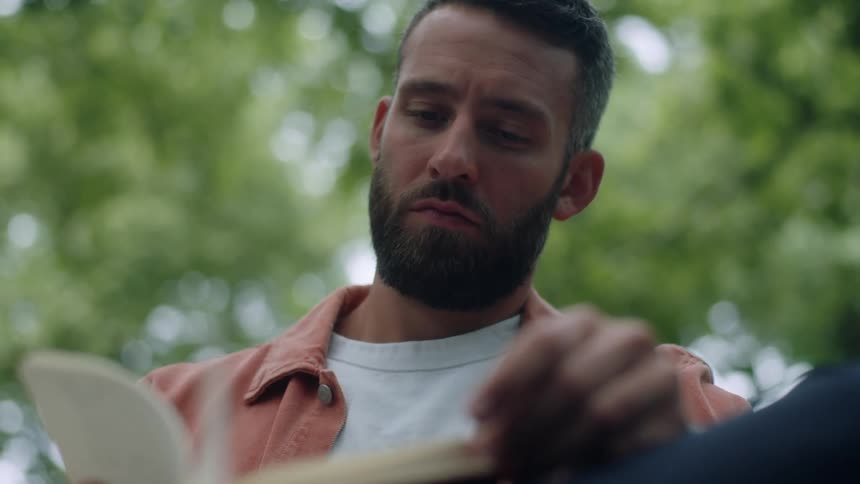 A man sits on a bench outdoors in a park reading a book