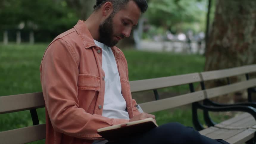 A man sits on a bench outdoors in a park writing on a notebook