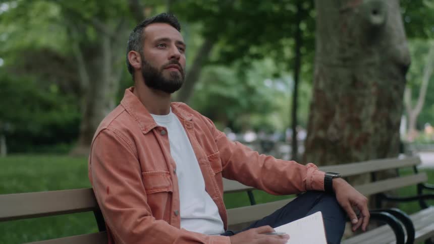 A man sits on a bench outdoors in a park writing on a notebook