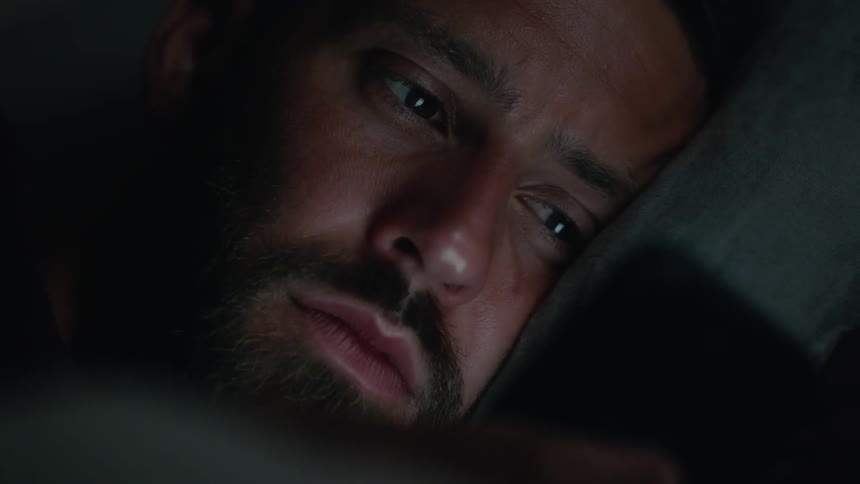 Close up of the face of a man resting in bed smiling while browsing his phone at night