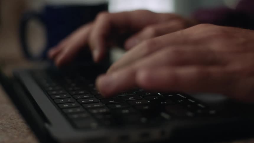 Person sits at a table with a coffee mug and types on a laptop