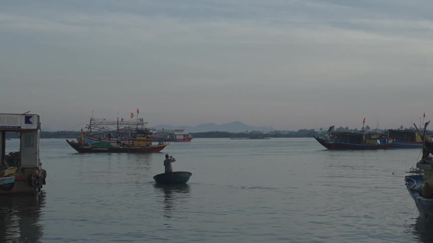 A person rowing on a small boat surrounded by several boats on an overcast morning