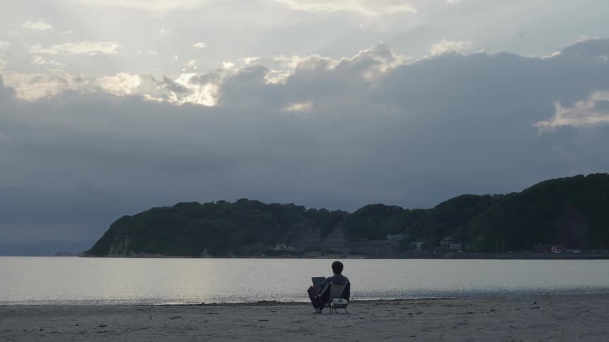 A solitary figure sits on a chair facing a beach with hills in the background under an overcast sky