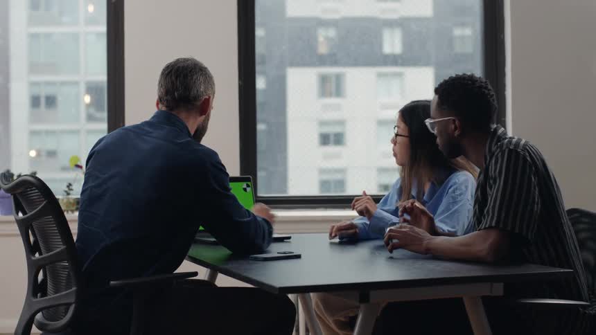Group of three people sitting at an office table with one of them using their laptop and the other two on their cell phone