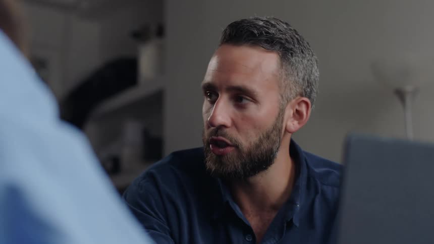 Portrait of a man sitting with a laptop in front of him talking to another person
