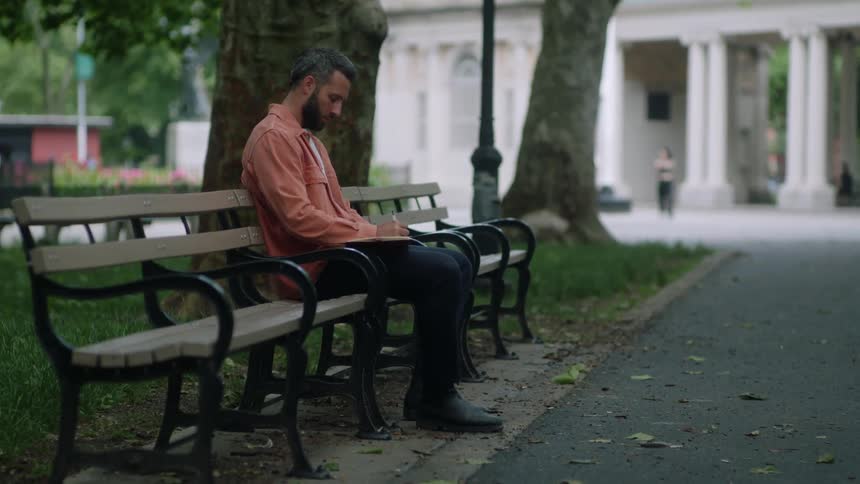 A man sits on a bench outdoors in a park writing on a notebook