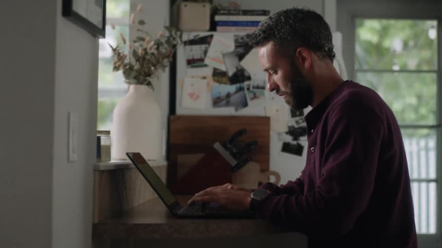 A man sits on a small table at home working on a laptop while looking at a phone