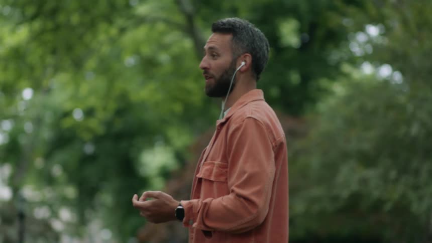 A man walks around a park speaking out loud while listening to an ear bud connected to a smartphone