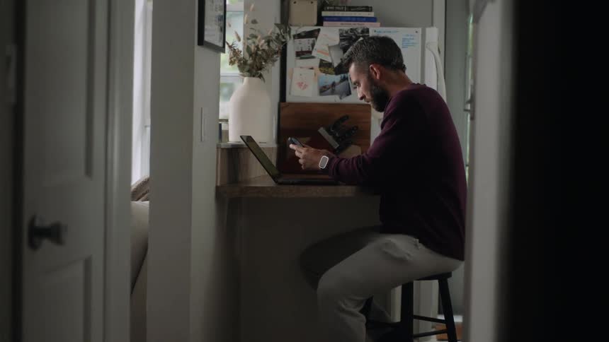 A bearded man sits at a small desk working on a laptop in a dimly lit room handling a smartphone