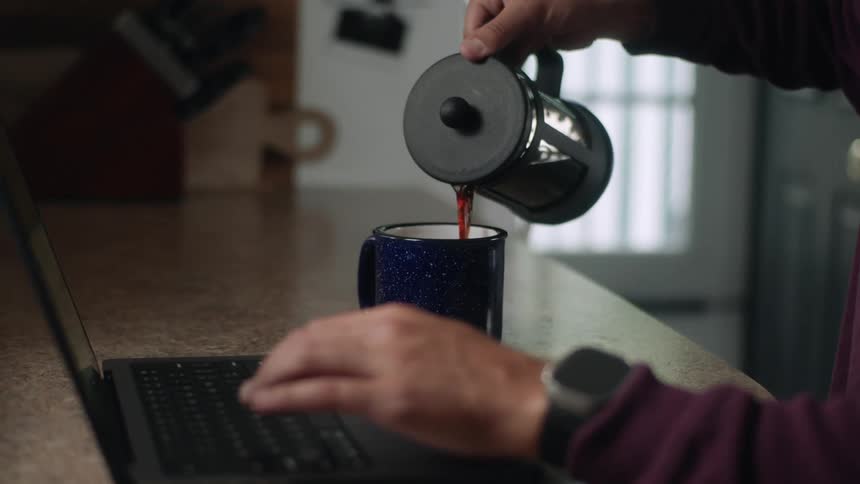 A man sitting on a desk at home serves himself a cup of coffee while working on a laptop