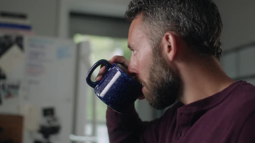 A man sitting on a table at home serves himself a cup of coffee while working on a laptop