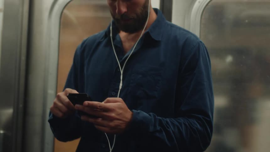 A man standing inside a subway train holding a smartphone connected to earphones