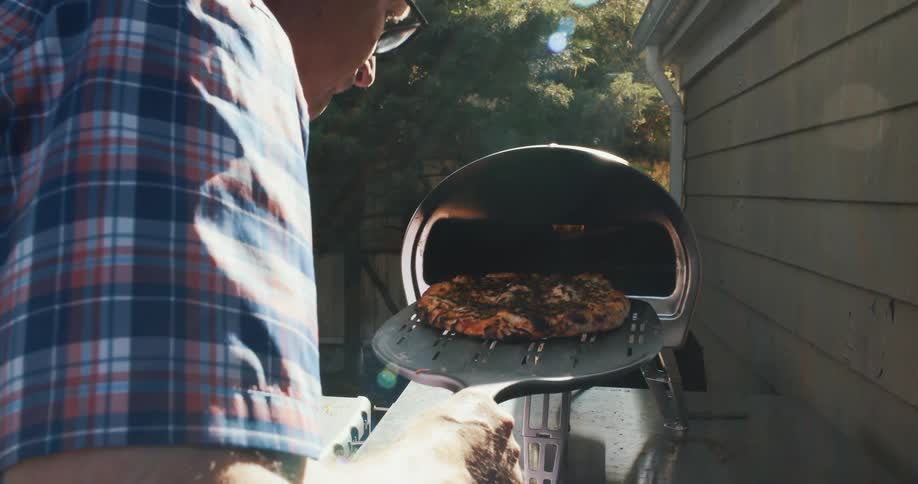 An older man pulls a homemade pizza out of an outdoor oven on a summer day