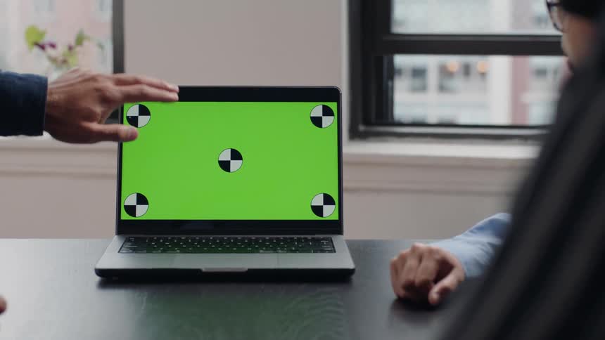 A laptop with a green screen and tracking markers sits on a table with business colleagues discussing it
