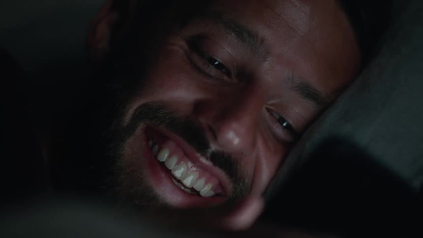 Portrait of a man lying down in a dark room using his cell phone smiling