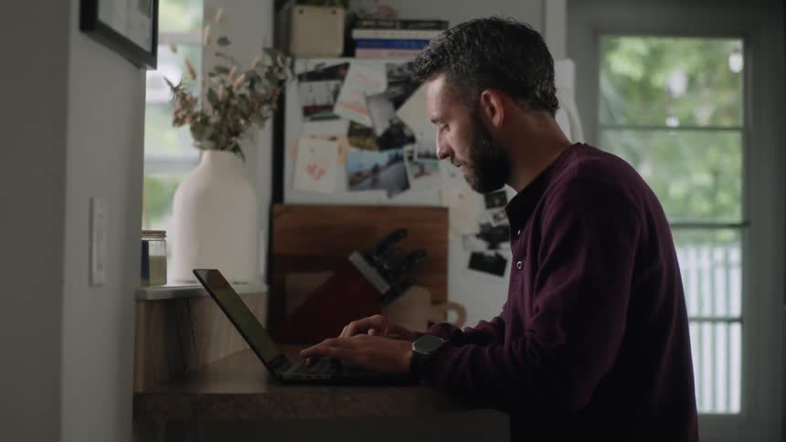 A bearded man sits at a small desk working on a laptop and checking his phone