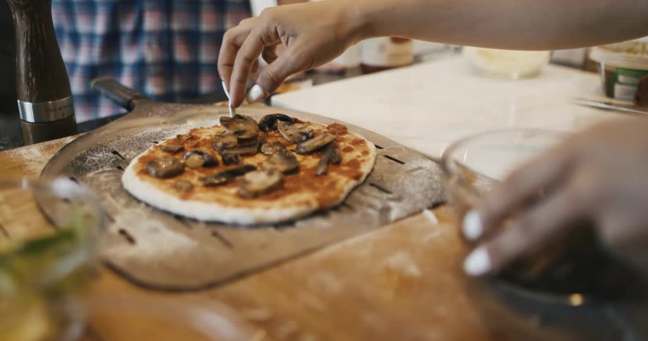 A hand arranges mushrooms over a freshly baked pizza on top of a wooding board