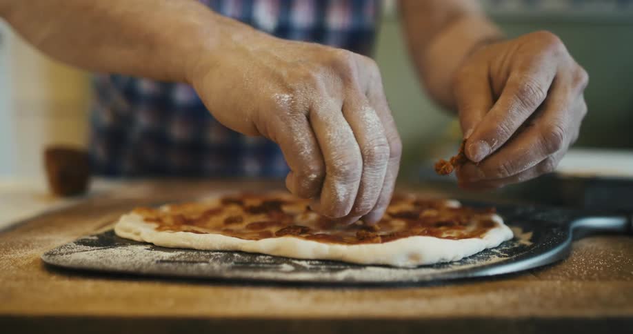 Man standing in the kitchen adds toppings onto a pizza