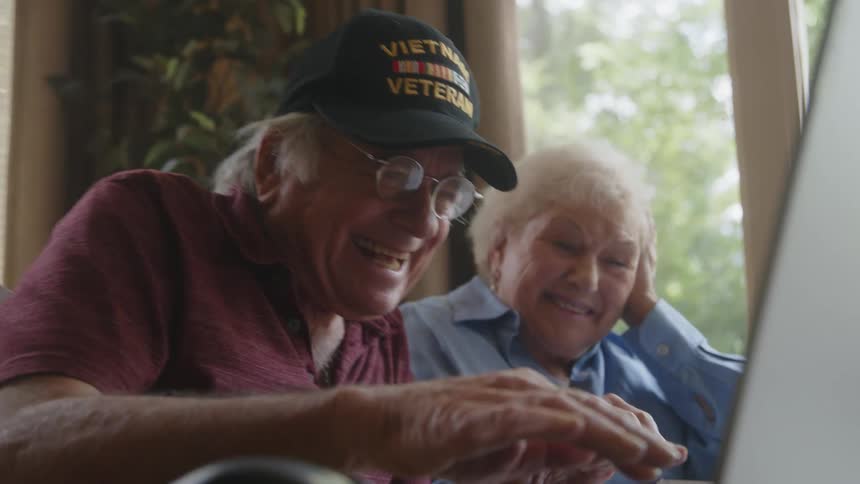 Smiling elderly couple sitting side by side with one of them typing