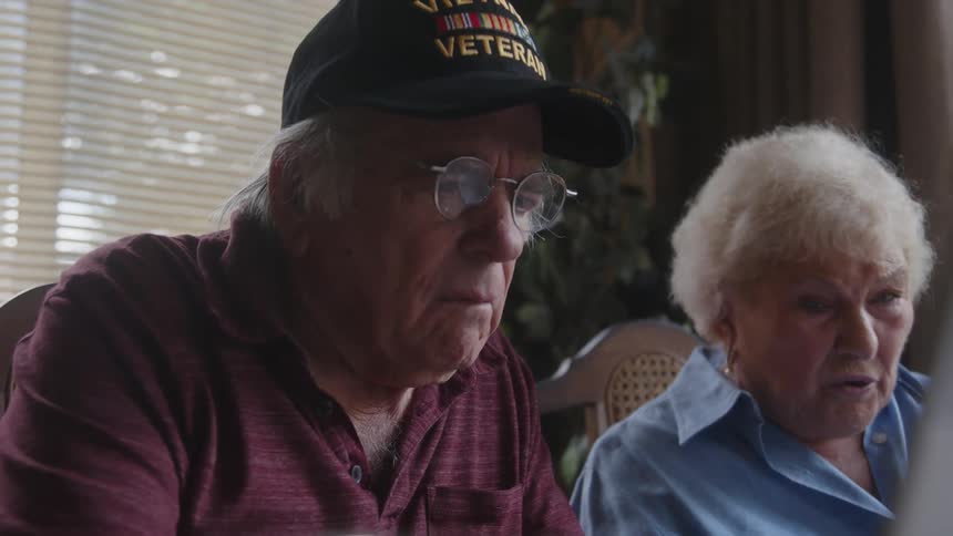 An elderly man looks at a laptop while his wife speaks angrily beside him
