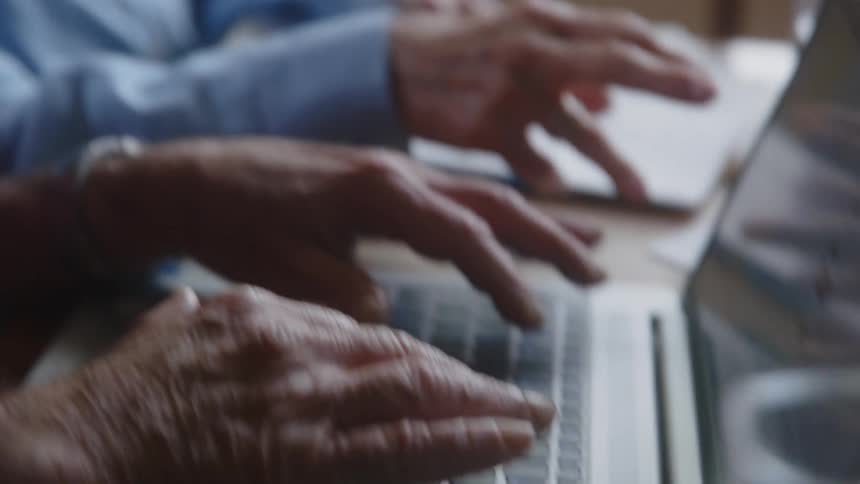 Two elderly people sitting at a table with one of them working on a laptop