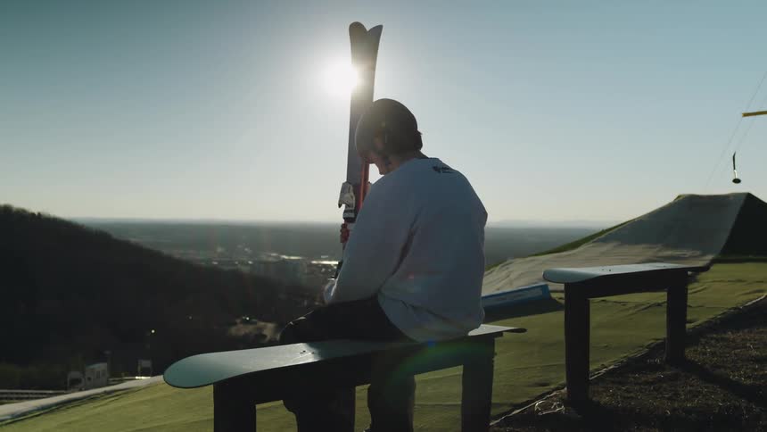 Man sitting on a bench outdoors with hills in the background and the sun low in the sky holding snow skiing equipment