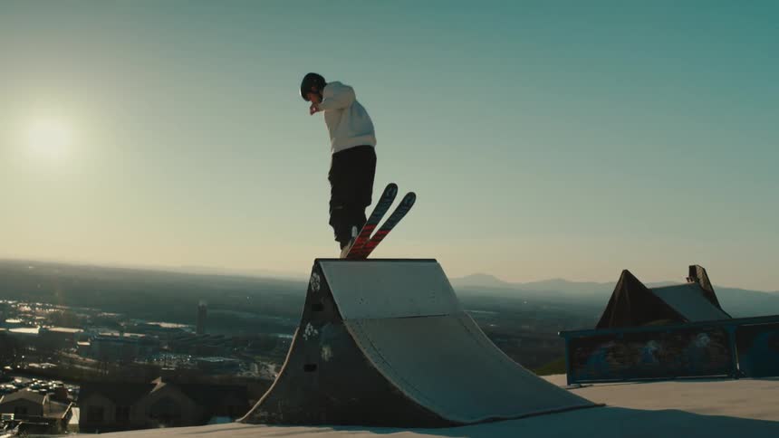 A young man skis over a ramp with a cityscape in the background
